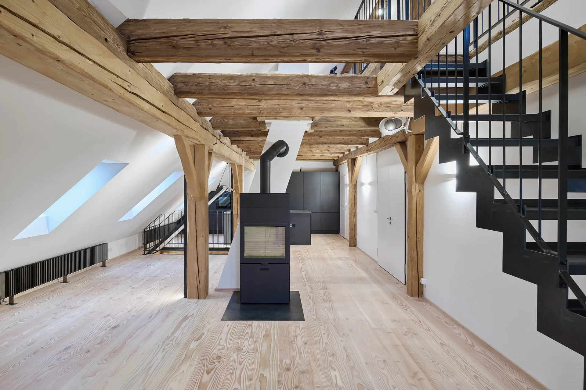 Husertal – Offener Wohnraum mit Dachstuhl, schwarzem Kaminofen und Stahltreppe